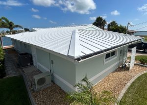 Single-story light blue house with a white metal roof, surrounded by gravel landscaping, palm trees, and a view of water in the background under a partly cloudy sky.