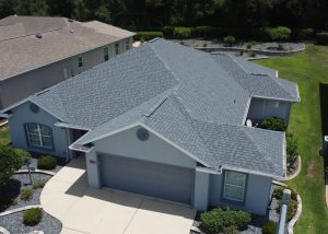 Aerial view of a single-story house with gray shingles, blue exterior walls, and a two-car garage, surrounded by shrubs and neighboring homes.