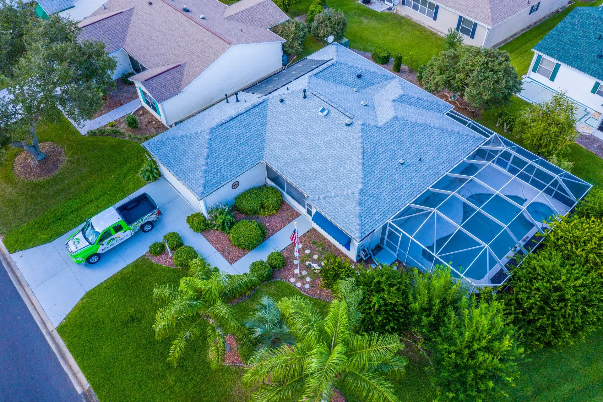 Aerial view of a suburban house with a screened-in backyard pool, green lawn, landscaping, and a green pickup truck parked in the driveway.
