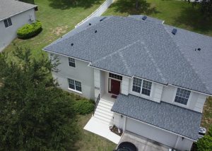 Aerial view of a two-story house with a gray shingle roof, white exterior, red front door, front steps, driveway, and surrounding green lawn.