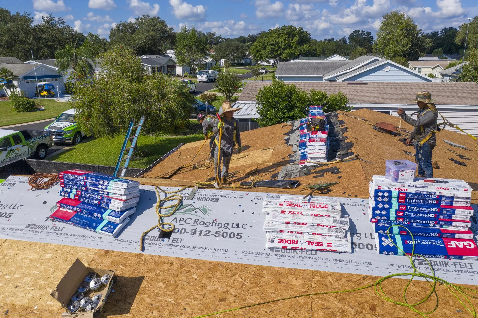Roofers work on installing shingles on a residential roof, with stacks of roofing materials and safety equipment visible on the rooftop.