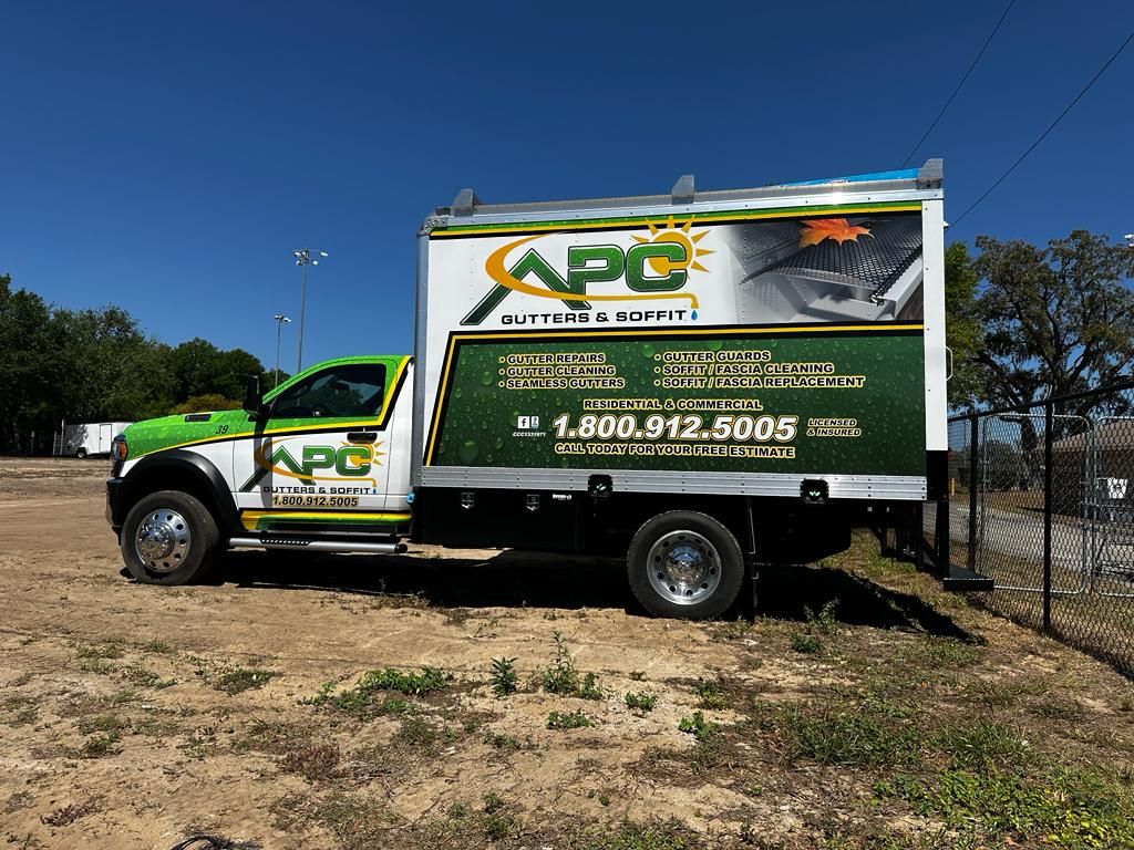 A truck with an APC Gutters & Soffit advertisement is parked on a dirt lot under a clear blue sky. The truck displays services and a contact phone number.