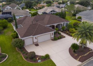 Aerial view of a single-story suburban house with a red roof, three-car garage, wide driveway, landscaped yard, and surrounding houses.
