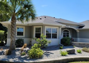 Single-story house with light gray exterior, white trim, and a dark roof, featuring a bay window, arched entryway, and landscaped front yard with gravel and various plants.