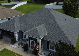 Aerial view of a gray, single-story house with a shingled roof, white trim, and a ladder propped against the porch; surrounded by green lawn and a white fence.