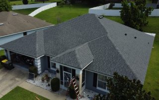 Aerial view of a gray, single-story house with a shingled roof, white trim, and a ladder propped against the porch; surrounded by green lawn and a white fence.
