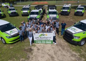 A group of people holding an APC Roofing LLC banner stands in front of several branded company trucks and vans in an outdoor area.