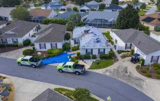 Aerial view of workers repairing the roof of a house in a suburban neighborhood, with two company trucks parked outside and materials on a blue tarp.