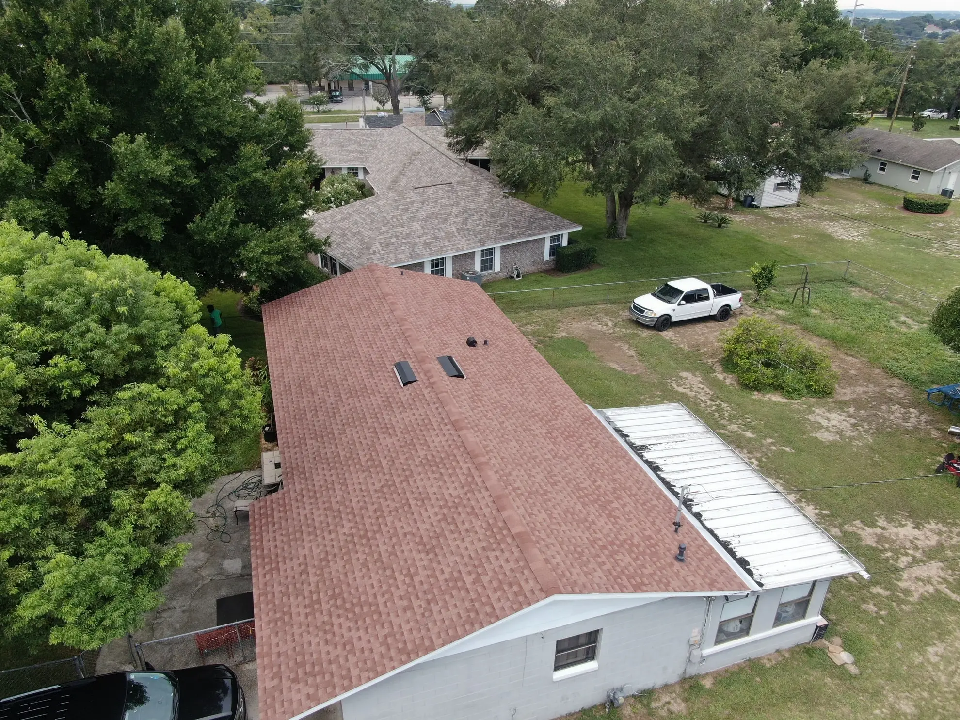 Aerial view of a house with a brown shingle roof, two skylights, a white truck parked nearby, trees, and other houses in the background.