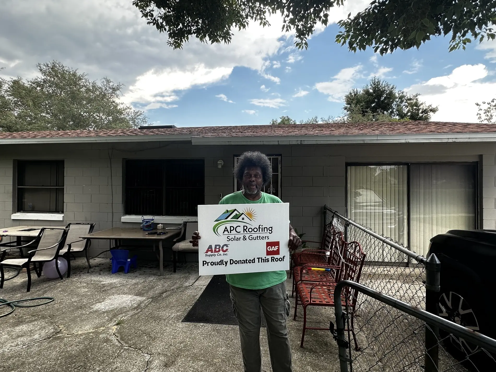 A person stands in front of a house holding a sign that reads, APC Roofing Solar & Gutters, Proudly Donated This Roof, with tables and chairs nearby.