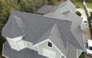 Aerial view of a light green house with gray shingle roofing, multiple gables, white trim, and a screened patio, surrounded by trees and a driveway.