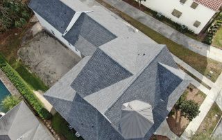 Aerial view of a house with a complex gray shingle roof and a covered patio area, surrounded by paved walkways and nearby buildings.
