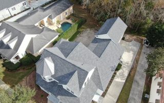 Aerial view of a residential property with gray roofs, a driveway, detached garage, and trees bordering the backyard. Neighboring house with a pool is visible.