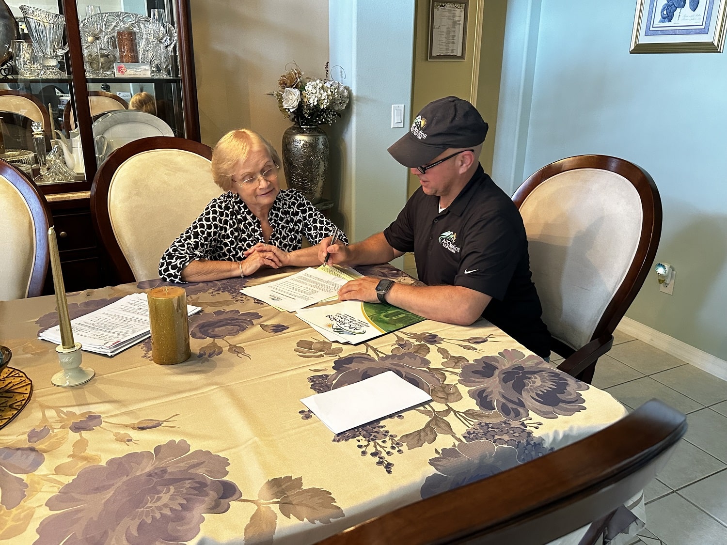 A roofing professional discusses paperwork and options with a homeowner inside her home, likely finalizing a roofing contract.